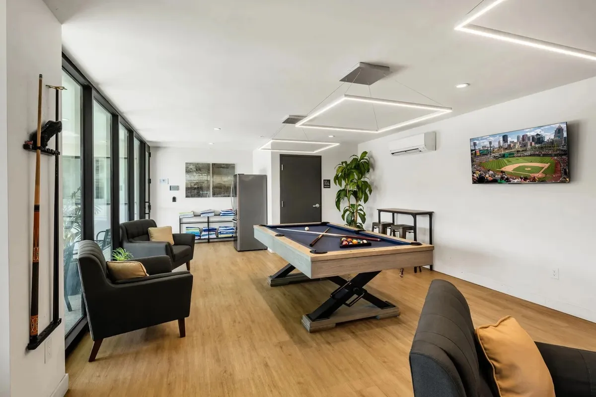 This interior shot captures the heart of the clubhouse, featuring a modern pool table, comfortable lounge seating, a large television screen, and lush potted plants. The casual and vibrant atmosphere invites guests to gather for friendly games or to relax and socialize in a resort-inspired environment.
