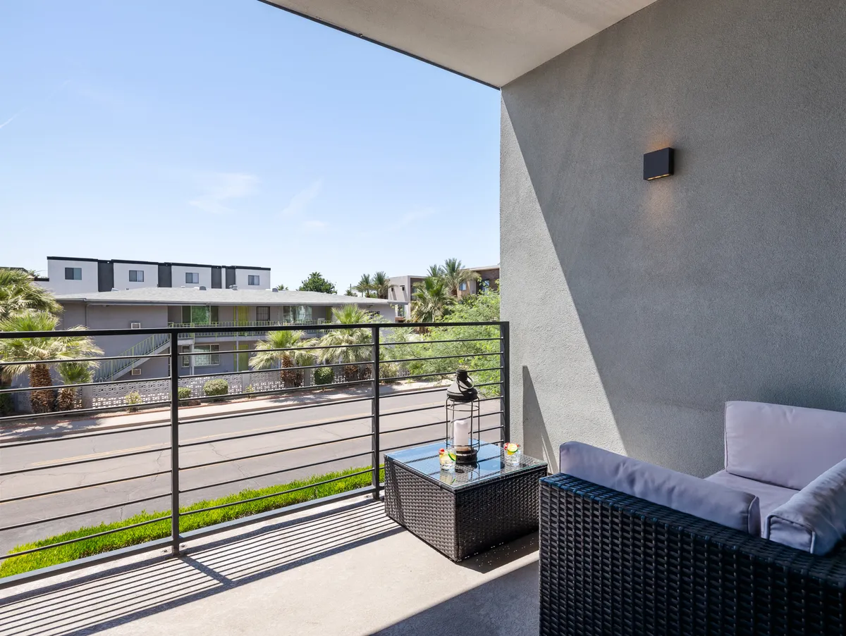A welcoming balcony space with cushioned seating, a small table adorned with decorative items, and a view of the street lined with palm trees. The atmosphere is tranquil, perfect for sipping coffee or sharing evening drinks while enjoying the Arizona skyline.
