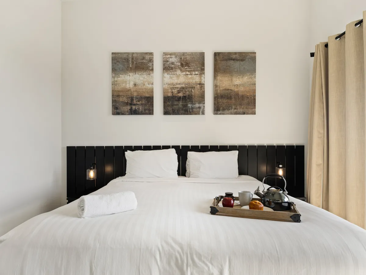 A cozy and inviting bedroom scene with a king bed dressed in soft white linens. A tray with beverages and snacks rests atop the bed, suggesting a peaceful morning ritual. Artwork decorates the wall, and the room is bathed in soft, natural light, creating a sense of relaxation and comfort.