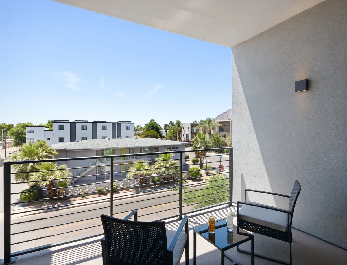 A peaceful balcony with comfortable seating overlooking palm trees and modern architecture, inviting guests to relax with a book or a drink while enjoying the sunset or morning sun. The elevated vantage provides a private outdoor escape connected to the bedroom.