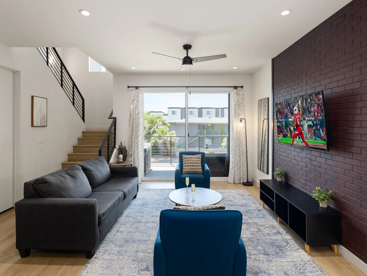 A cozy living room featuring a comfortable sofa, a blue armchair, a coffee table, and a large television set against a textured brick wall. The layout is ideal for relaxing, streaming shows, or spending quiet moments with loved ones.