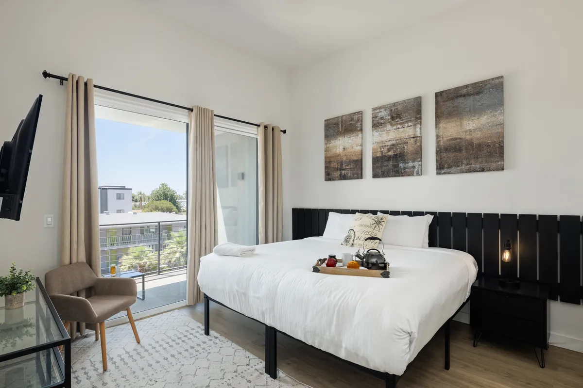 A serene king bedroom featuring crisp white linens, tasteful artwork on the wall above the headboard, and a sliding glass door that opens to a private balcony. The natural light floods the space, emphasizing the calm and inviting atmosphere. A Smart TV is mounted for easy viewing from the plush bed.