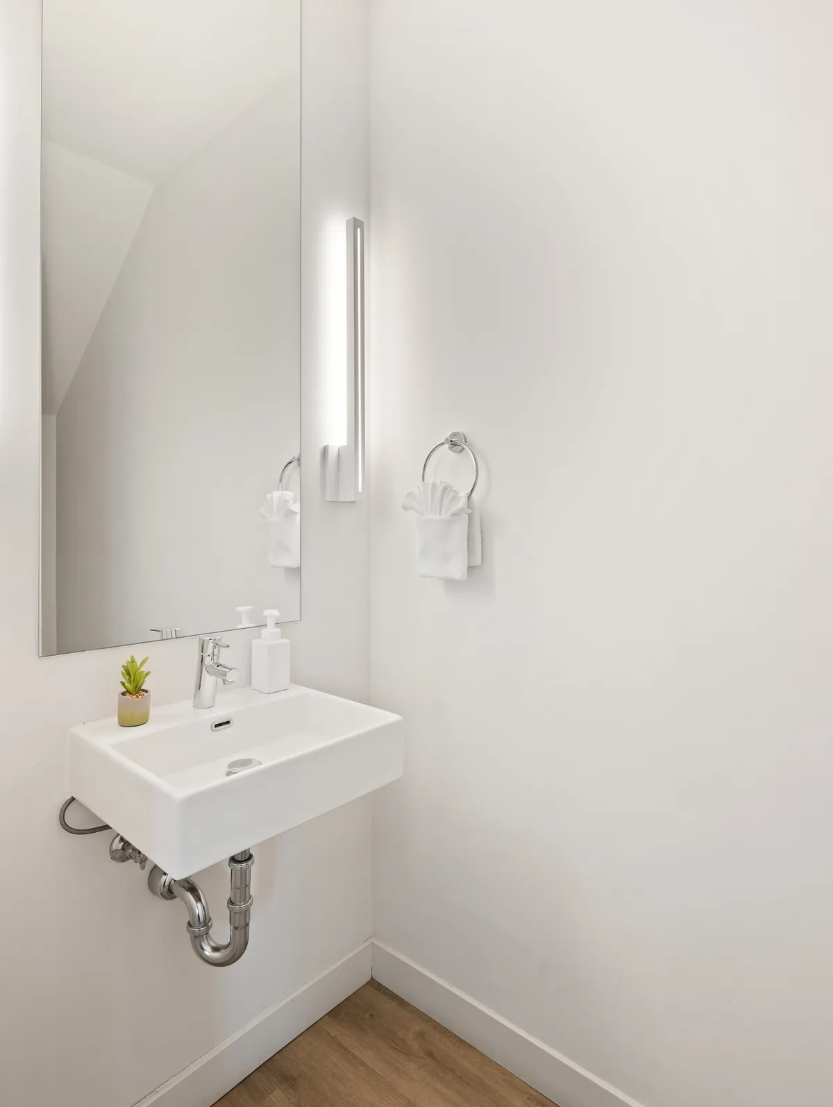 A serene and minimalist bathroom with a sleek sink, a round mirror, a small plant, and a neatly hung towel. The ambiance is calming and uncluttered.